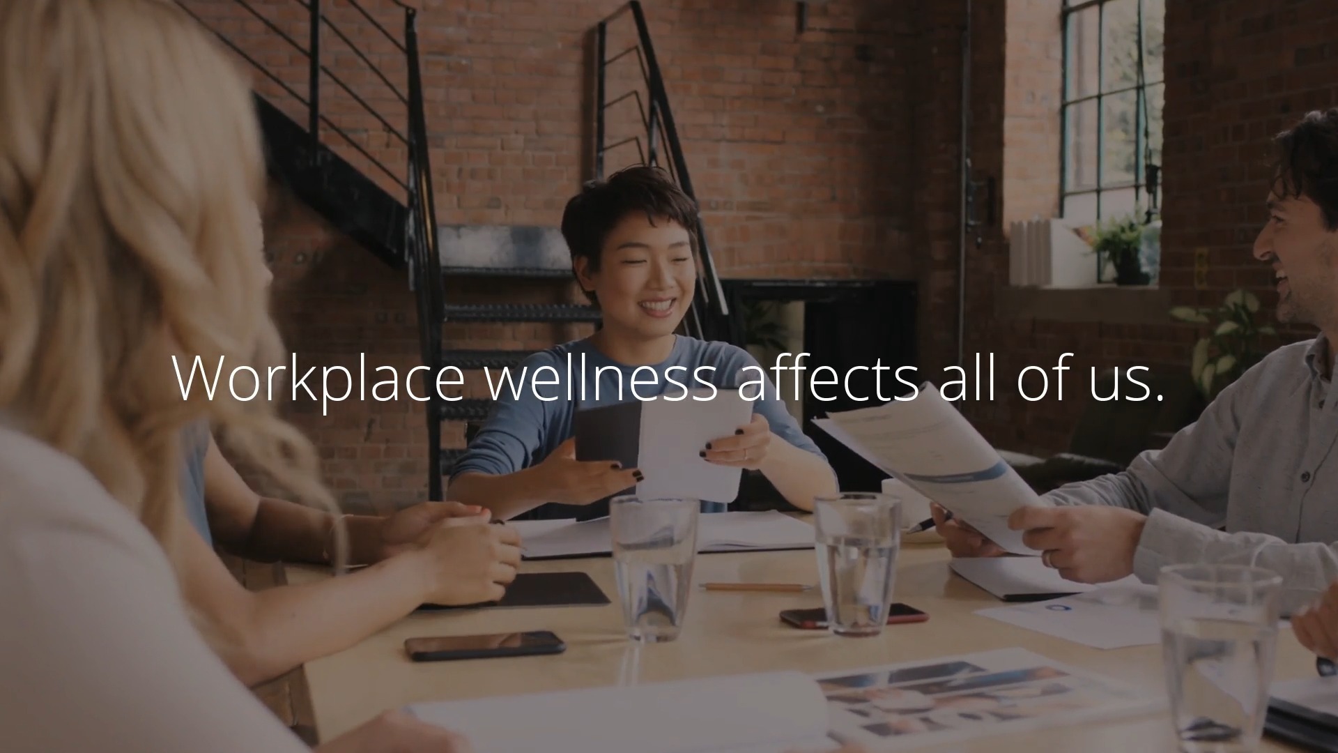 Group of professionals discussing documents at a meeting table with a Biteable video maker presentation in the background displaying the message, "workplace wellness affects all of us.