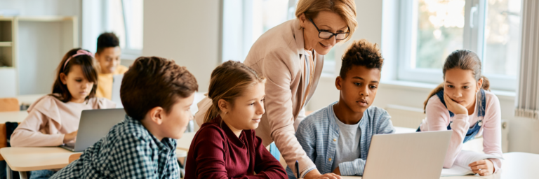 Teacher assisting diverse group of elementary students using a laptop in a classroom.