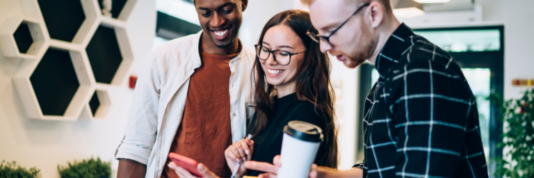 Three colleagues, one Black man and two white individuals, a woman and a man, look at a smartphone and smile together in a modern office setting.