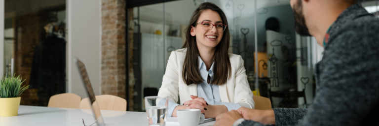 Woman in glasses and white blazer smiling during a meeting with a male colleague in a modern office setting.